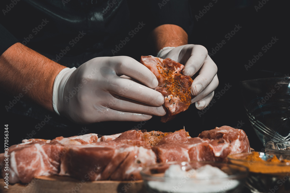 A man in gloves prepares and marinates pork with spices for cooking kebabs, uses salt, curry and basil. - obrazy, fototapety, plakaty 