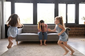 Young tired single mother suffers from headache closed eyes touch forehead sitting on couch while her daughters running around her and shouting, female babysitter feels exhausted by noisy kids