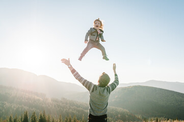 Dad playing with daughter and throws her high in the sky in mountains at the sunset time. Concept of friendly family. Portrait Dad and child enjoying autumn vacation. A place for text, advertising.