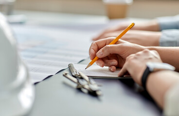 construction business, architecture and buiding concept - close up of architect with blueprint, notebook and pencil working at office