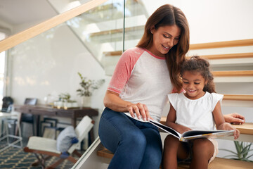 Hispanic Mother And Daughter Sitting On Staircase In Modern Home Reading Book Together