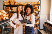 Two Beautiful Young Bakers At The Bakery. Shot Of A Young Woman Showing Her Colleague Something On Her Clipboard While They Stand In Their Bakery Shop. Smiling Young Bakers At The Bakery Shop