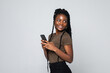 Typing message to friend. Confident young African woman holding mobile phone and looking at it with smile while standing against grey background