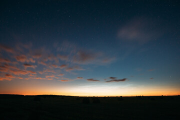 Wall Mural - Night Starry Sky With Glowing Stars Above Countryside Landscape. Light Cloudiness Overcast Above Rural Field Meadow In Early Spring. Illumination Lights Of Town On Horizon