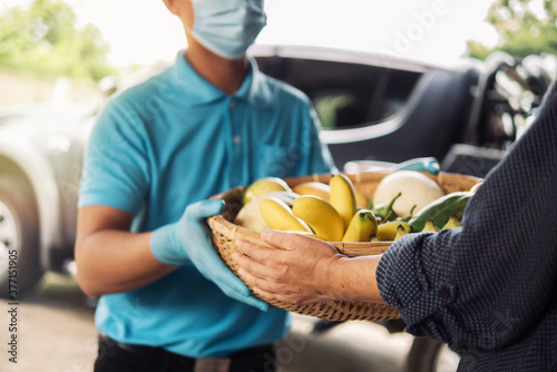 Senior woman hand receiving fresh fruits and vegetables in bamboo basket from delivery man wearing protective gloves and face mask. Food delivery in COVID-19 pandemic and senior people social care.