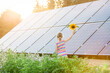Young 6 year old blonde girl child standing in front of small solar panel farm in countryside. Renewable energy concept. Bright yellow warm sun lens flare.