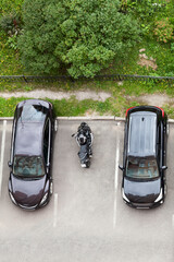 Motorcycle is parked on vehicle parking space between two passenger cars. Top view, vertical