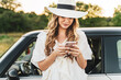 © Miljan Živković - Front view on beautiful caucasian woman using mobile phone texting while standing by her car in nature in bright summer or autumn day - travel and fall concept yellow filter