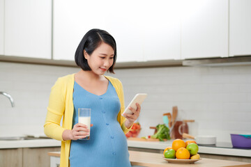 Young pregnant woman holding glass of milk near table with food in kitchen