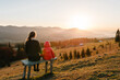 © Serhii - Happy family: mom and daughter, enjoying time together, sitting on sunset on top of foggy mountain. Tourists on background view mockup. Hikers looking on sunlight in trip in country Europe. Back view.