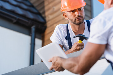 Wall Mural - Selective focus of builder with hammer looking at colleague with digital tablet on roof of building