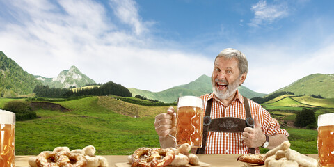 Wall Mural - The senior happy smiling man with beer dressed in traditional Austrian or Bavarian costume holding mug of beer, mountains on background, flyer ready for ad. The celebration, oktoberfest, festival