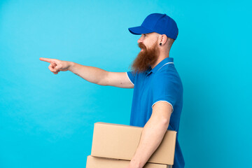 Redhead delivery man over isolated blue background pointing to the side to present a product