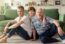 Portrait Of Three Generations Of Men Sitting On The Warm Floor In Living Room Hugging. Cute Little Boy With Young Dad And Grandfather Enjoy Family Leisure Weekend Together
