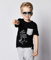 Wall Mural - Cool blond kid boy in sunglasses, black t-shirt with dinosaur print and gray pants pointing finger at camera 