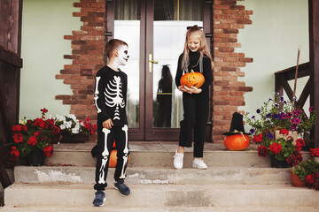 Brother and sister celebrate halloween on the street near the house in costumes and make-up with a pumpkin lantern, play and laugh