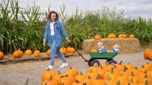 Young Mom Pulling Her Twin Babies In A Wagon In The Pumpkin Patch. Happy Family Concept