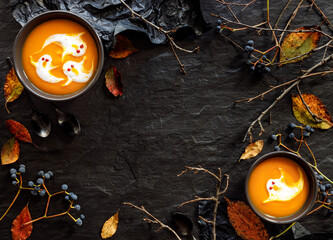 Halloween background with pumpkin soup decorated with cream ghosts, autumnal leaves on a black background  with copy space