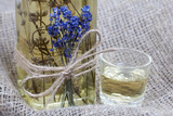 Fototapeta Storczyk - A bottle of lavender tincture. A bouquet of lavender is tied to the bottle. Nearby is a glass of drink. On coarse linen. Close-up shot.