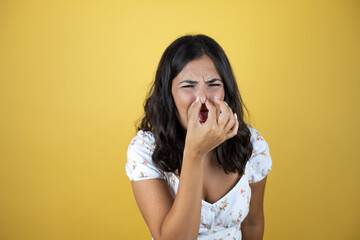 Wall Mural - Beautiful woman over yellow background smelling something stinky and disgusting, intolerable smell, holding breath with fingers on nose