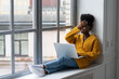 Happy African American woman freelancer with afro hairstyle wear yellow cardigan sitting on windowsill, working on laptop, talking in video chat, laughing with toothy smile. Forgot, bad memory concept