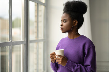 Pensive African American millennial young woman in trendy jewelry big earrings wear purple sweater thinking, looking through the window, holding white mug, drinking coffee or tea. 