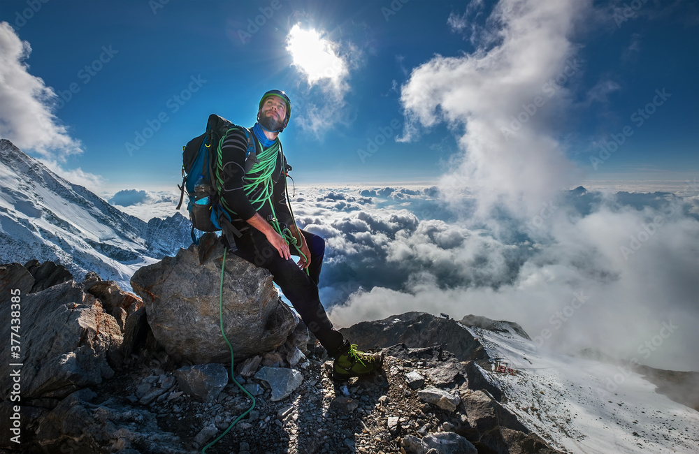Climber in a safety harness, helmet, and high mountaineer boots with picturesque clouds background sitting at 3600m altitude on big rock and looking at summit during Mont Blanc ascending, France route - obrazy, fototapety, plakaty 