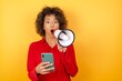 © Roquillo - Young arab woman with curly hair wearing red shirt holding a megaphone on yellow background opens mouth hold telephone reading advert unbelievable big shopping prices