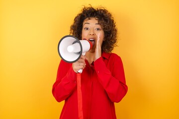 Wall Mural - Young arab woman with curly hair wearing red shirt holding a megaphone on yellow background, shouting excited to front.