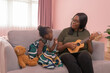 © tampatra - Portrait of smiling business black African american woman, a mom with her daughter playing an ukulele music in family relationship concept on pink background. A black kid toddler girl with her parent.