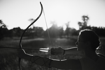 Young Caucasian female archer shooting with a bow in a field at sunset.