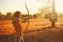 Young Caucasian Female Archer Shooting With A Bow In A Field At Sunset.