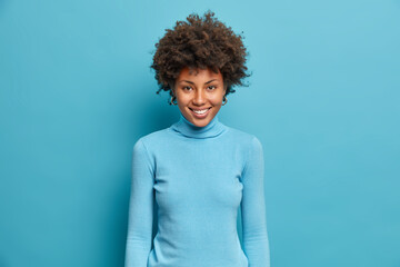 Wall Mural - Shot of attractive cheerful Afro American woman rejoices nice day and smiles broadly, wears casual blue turtleneck. Vivacious teenage lady has perfect white teeth, expresses positive emotions