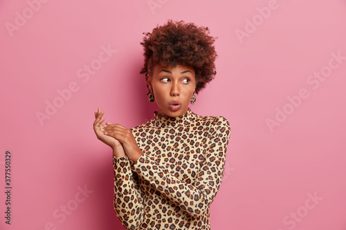 Fotografía Young stylish pretty woman with Afro hair looks surprisingly aside, keeps mouth opened and rubs hands