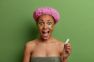 Wall Mural - Isolated shot of crazy surprised dark skinned female model with opened mouth holds tampon for protection during menses, enjoys beauty treatments after taking shower and poses against green background.