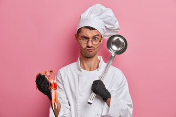 Displeased professional angry Caucasian man chef holds uncooked crayfish and ladle, prepares gourmet tasty dinner. Cook with kitchenware cooks seafood dish, works in restaurant. Cooking concept