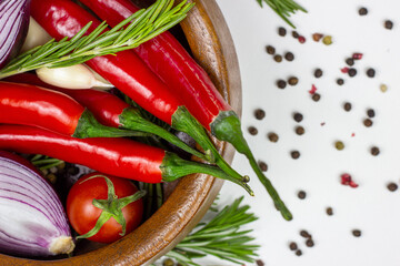 Wall Mural - Top view of summer vegetables: onion, garlic, tomato, red chili pepper, rosemary in brown wooden bowl and peppercorns on white table background.