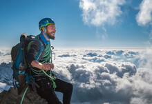 Smiling Bearded Climber In A Safety Harness, Helmet, And On Body Wrapped Climbing Rope Sitting At 3600m Altitude On A Cliff And Looking At  Picturesque Clouds During Mont Blanc Ascending, France Route