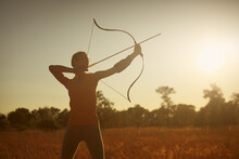 Young Caucasian Female Archer Shooting With A Bow In A Field At Sunset.