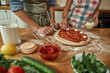 © Svitlana - Add cheese. Cropped shot of couple making pizza together at home. Man in apron adding, applying tomato sauce on the dough while woman adding mozzarella cheese. Hobby, lifestyle