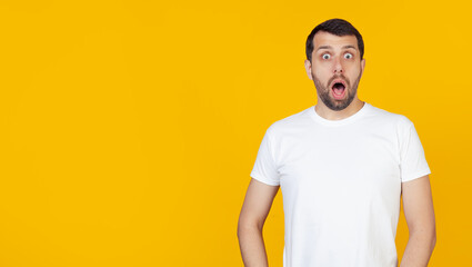 Young man with beard in white t-shirt afraid and shocked by unexpected expression, fear and excited face, stands against isolated yellow background