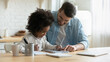 Smiling young european man helping little adopted african american daughter with homework, sitting together at table. Happy small biracial child girl involved in doing school tasks with foster father.