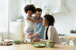 © fizkes - Smiling young european man holding on hands pretty small biracial adopted girl daughter, watching happy african american mother preparing healthy food, chopping fresh vegetables for salad in kitchen.