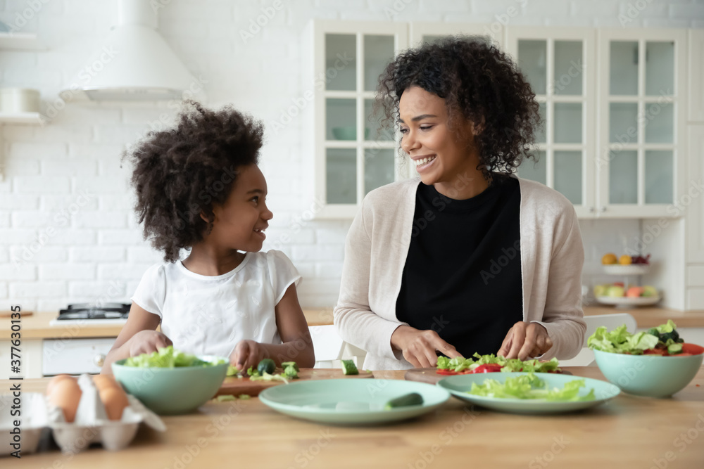 Happy small african american kid girl communicating with smiling biracial mother while preparing salad in modern kitchen, playful multiracial family having fun, enjoying cooking together at weekend. - obrazy, fototapety, plakaty 
