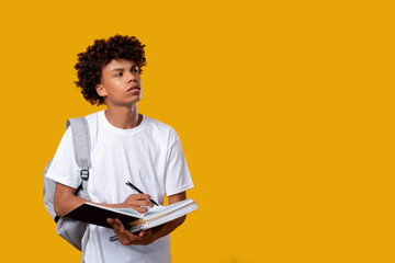 Doubtful black teenager. Think idea. Puzzled african male student writing in notebook isolated on orange copy space. Educational task. Find decision. Knowledge and skills