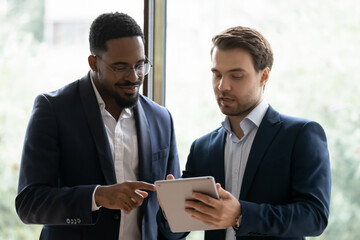 Wall Mural - Multiethnic businessmen look at tablet screen discuss business project together. Smiling diverse multiracial male colleagues brainstorm cooperate on pad in office. Technology, teamwork concept.