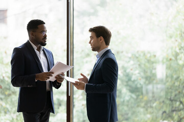 Wall Mural - Serious multiracial businessmen stand in office boardroom brainstorm discuss financial paperwork. Focused multiethnic diverse male colleagues talk analyze company paper document. Cooperation concept.