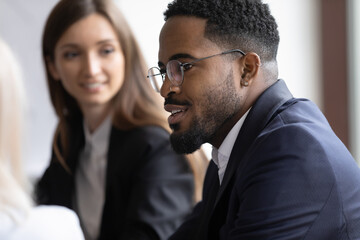 Wall Mural - Close up of confident young African American male employee in glasses brainstorm at team meeting with colleagues. Biracial businessman talk discuss idea with multiracial coworkers at office briefing.