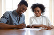 © fizkes - Excited African American young couple sit at desk put signature on contract close deal with realtor or broker, happy biracial husband and wife sign document agreement buying first house together