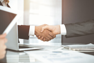 Unknown diverse business people are shaking hands finishing contract signing in sunny office, close-up. Business handshake concept